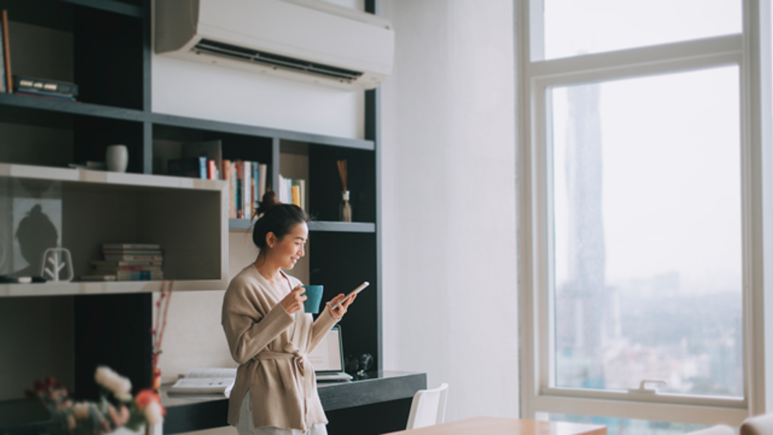 Woman at home drinking coffee and adjusting the temperature of her air conditioner on her smartphone
